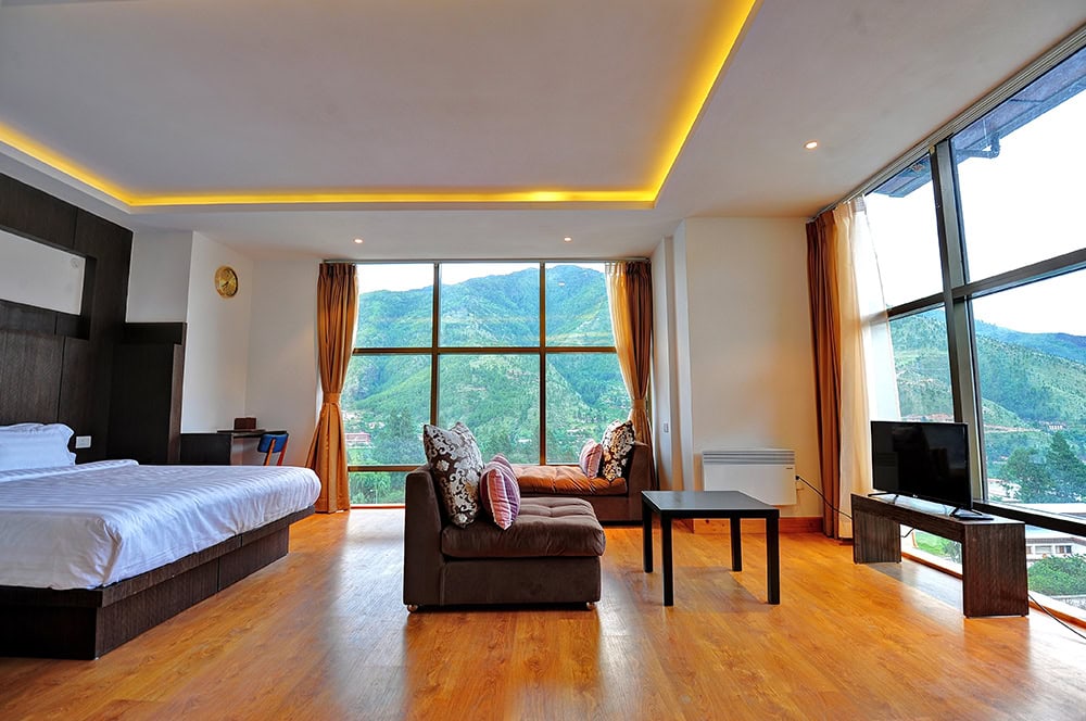 A spacious modern hotel room with a bed, sofa, coffee table, TV, and large floor-to-ceiling windows offering a scenic view of green mountains and a valley outside. Warm lighting and wood flooring add a cozy feel.