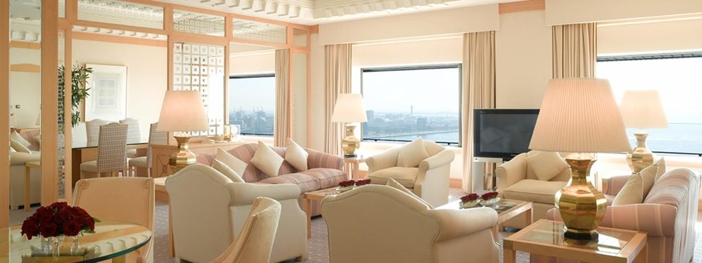 Spacious, elegant living room with cream-colored sofas and armchairs, large windows with city and water views, light wood furniture, mirrored walls, and table lamps with pleated shades.