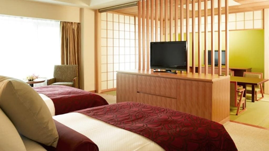 A modern hotel room with two beds, a wooden partition, a TV on a cabinet, a sitting area by the window, and a dining area with Japanese-style decor and sliding paper doors.