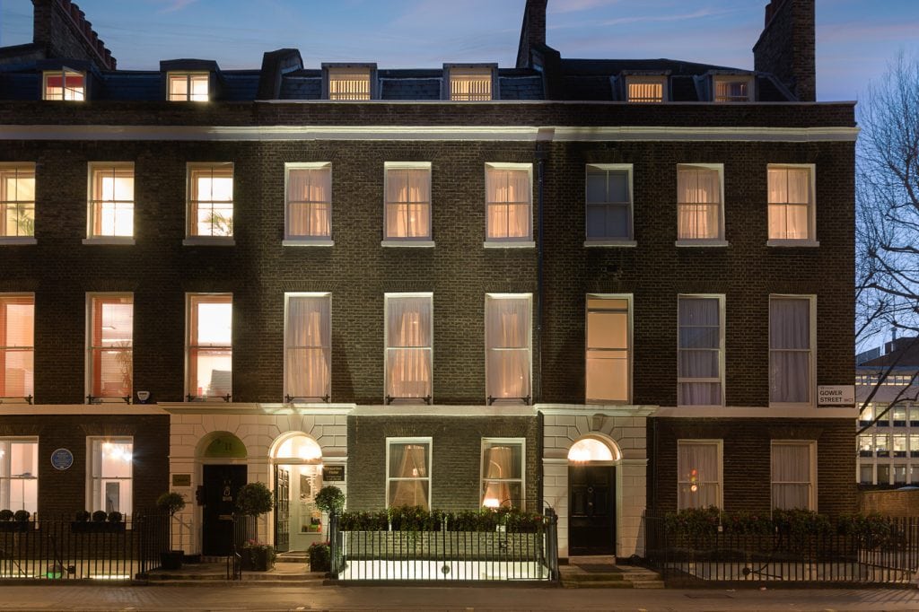 A row of dark brick townhouses at dusk, warmly lit from inside. Tall windows, black doors, white trim, and iron railings line the front. Some trees are visible on the right side.