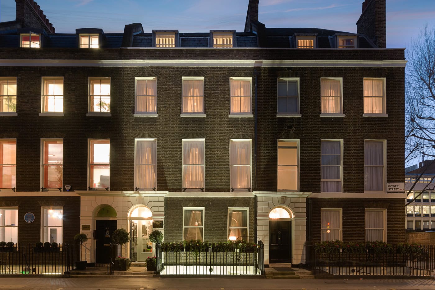 A row of dark brick townhouses at dusk, warmly lit from inside. Tall windows, black doors, white trim, and iron railings line the front. Some trees are visible on the right side.