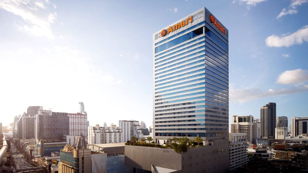 A tall modern skyscraper with blue-tinted windows and the orange AIA logo on top, surrounded by other buildings in a cityscape under a partly cloudy sky.