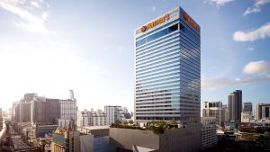A tall modern skyscraper with blue-tinted windows and the orange AIA logo on top, surrounded by other buildings in a cityscape under a partly cloudy sky.