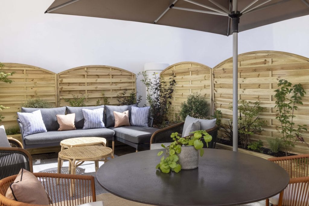 A cozy outdoor patio with a gray sectional sofa, cushioned chairs, and a round table with a potted plant, set under a large umbrella. Wooden privacy screens and green plants decorate the space.