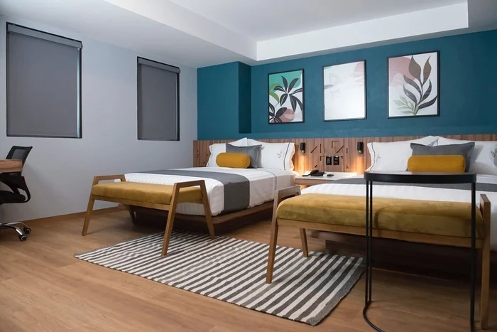 Modern hotel room with two double beds, mustard yellow benches, a striped rug, a desk with a chair, and three framed abstract artworks on a teal accent wall. The window shades are drawn.