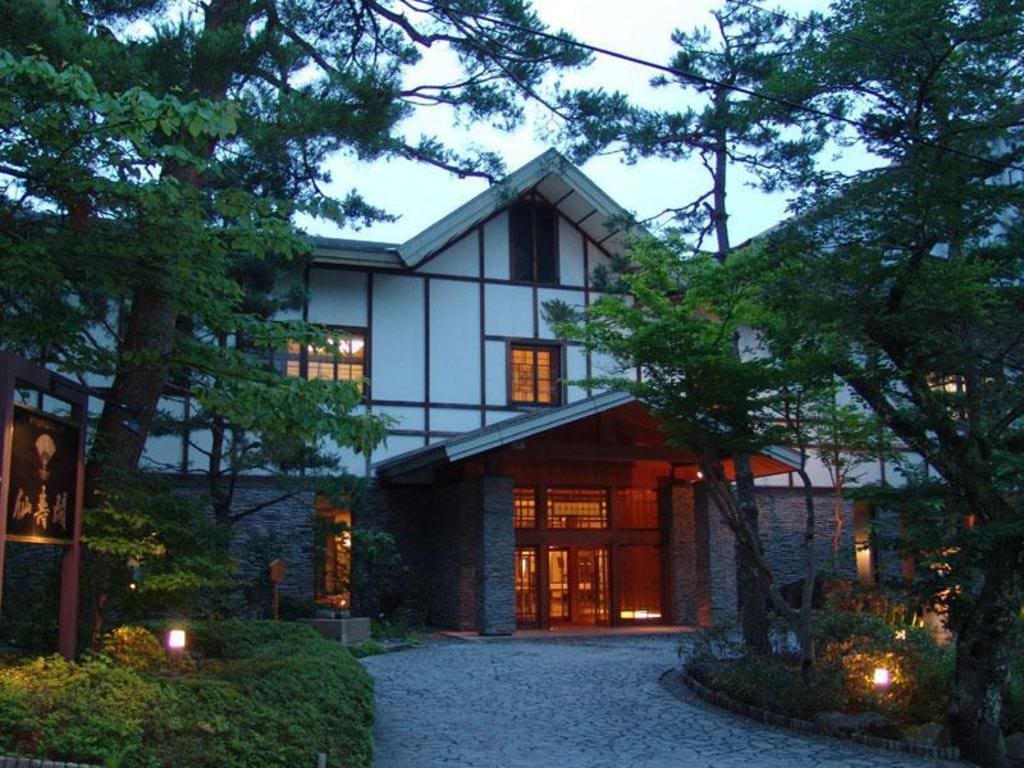 Traditional Japanese-style building surrounded by lush green trees, with warm lights glowing from windows and along a stone pathway leading to the entrance at dusk.