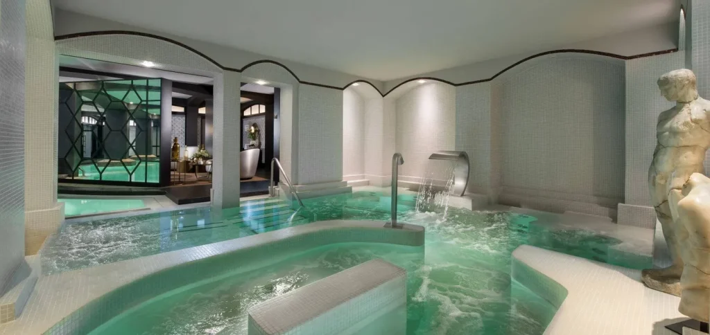 Luxurious indoor spa with glowing turquoise pools, waterfall feature, curved white tile walls, a classical statue, and elegant seating area in the background. Relaxing, serene atmosphere.
