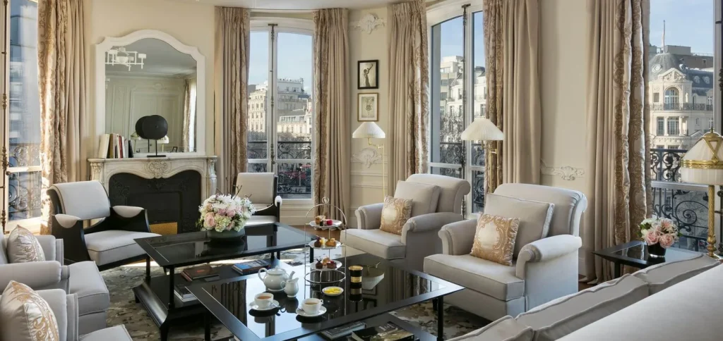 Elegant living room with large windows, cream-colored walls and curtains, plush armchairs, a black fireplace, and a coffee table set with tea and pastries. Parisian buildings are visible outside. Flowers decorate the room.