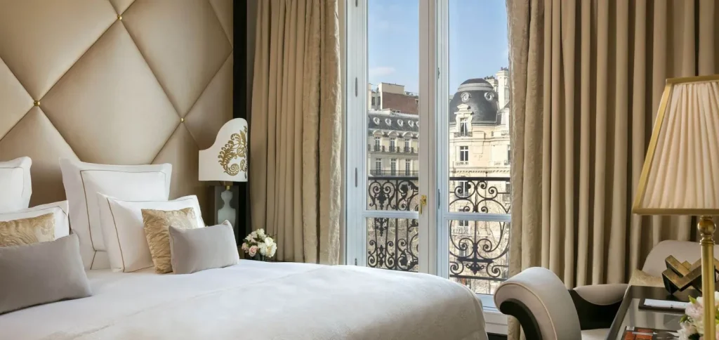 A luxurious hotel room with a white bed, gold and beige decor, large windows with beige curtains, and a view of ornate Parisian buildings outside. A lamp, chair, and flowers are visible in the elegant setting.