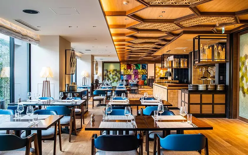 A modern, stylish restaurant with wooden floors, blue chairs, set tables with glassware, and a patterned wooden ceiling. Large windows provide natural light and colorful artwork decorates the far wall.