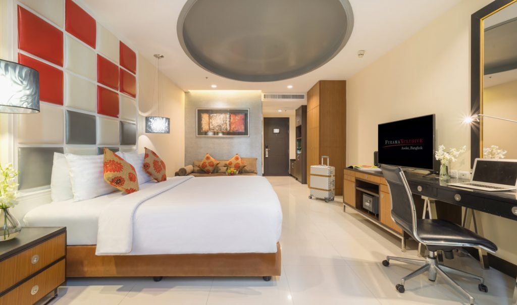 A modern hotel room with a large bed, colorful pillows, abstract wall art, a desk with a laptop, flat-screen TV, and a sitting area with a sofa. The room features bright lighting and stylish decor.