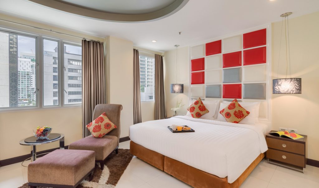 A modern hotel room with a large bed, red and gray accent headboard, two nightstands, an armchair with an ottoman, a round table with fruit, and windows overlooking city buildings.