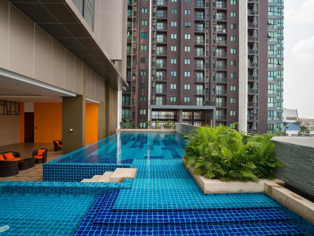 Outdoor swimming pool with blue tiles beside a modern high-rise apartment building, featuring green plants and orange seating in a shaded lounge area.