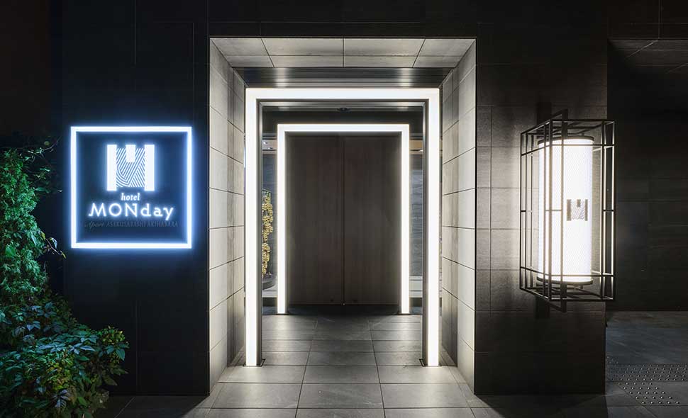 The entrance of hotel MONday is lit with white LED lights, featuring a modern black and gray facade, a glowing sign displaying the hotel name, and a large wooden door at the center.