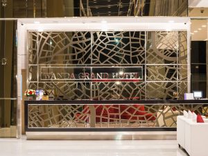 Hotel reception desk with a modern, geometric mirrored wall behind it, featuring the words WANDA GRAND HOTEL. A vase of flowers, a computer, and a white sofa with red pillows are visible in the contemporary lobby.