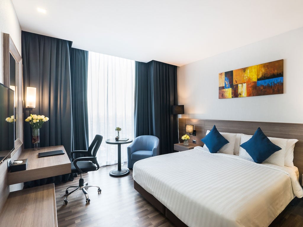 A modern hotel room with a large double bed, blue accent pillows, a desk with chair, small round table with two chairs, floor-to-ceiling window with dark curtains, and abstract art on the wall.