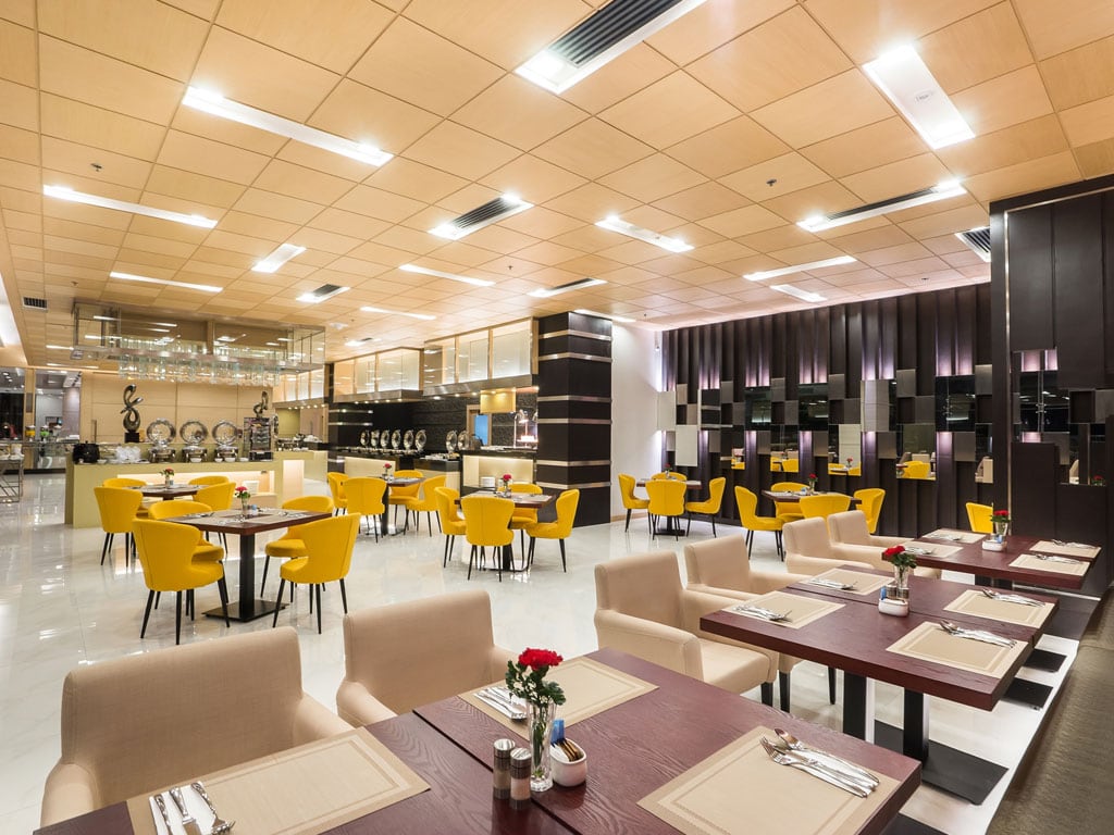 Spacious modern restaurant with beige and yellow chairs, wooden tables set with placemats, and decorative red roses. Buffet stations line the back wall under warm ceiling lights.