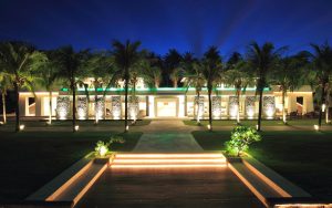 A modern, well-lit building with large windows is surrounded by palm trees at night. Illuminated pathways and stairs lead to the entrance, creating a warm and inviting atmosphere.