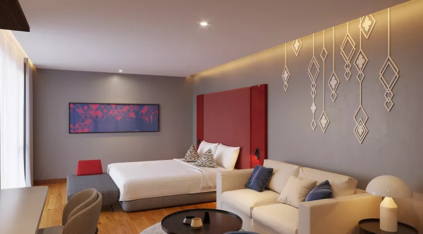 A modern hotel room with a double bed, red headboard, decorative wall art, a white sofa with cushions, a round coffee table, and wooden flooring. Large windows on the left let in natural light.