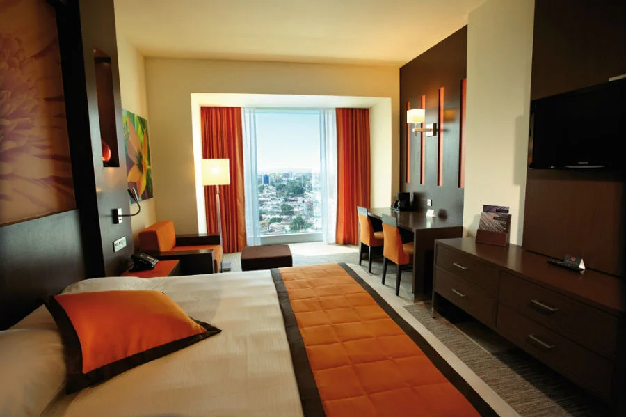 Modern hotel room with a large bed, orange accents, a desk, a TV, and floor-to-ceiling windows with red curtains, offering a city view and plenty of natural light.