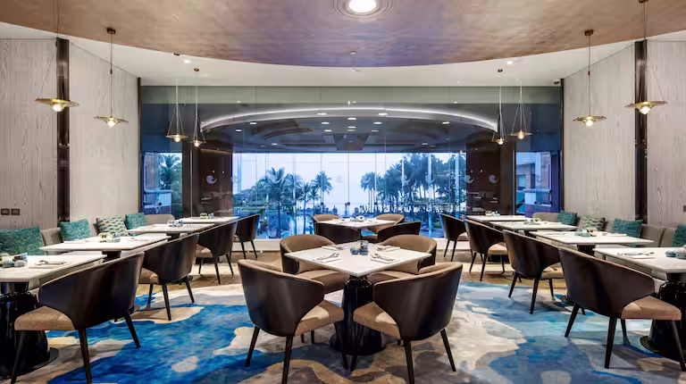 Modern restaurant interior with round tables, cushioned chairs, blue-patterned rug, and pendant lights. Large glass windows at the back reveal palm trees and a view of the ocean.
