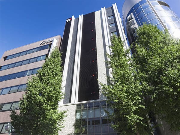 A modern multi-story office building with large vertical black and white accents and glass windows, flanked by green leafy trees under a clear blue sky. Another building with a KD logo is visible on the left.