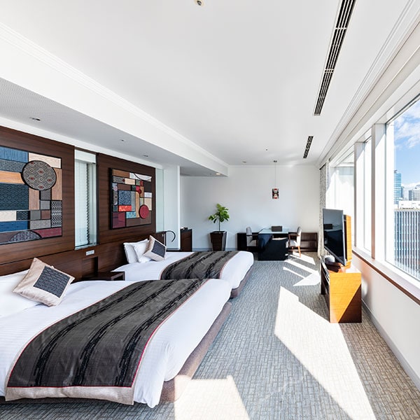 Modern hotel room with two double beds, colorful wall art, a seating area near large windows, a TV on a wooden stand, and city views with bright natural light filling the space.