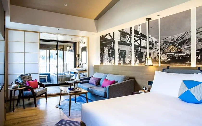 Modern hotel room with a large bed, gray sofa, coffee table, armchair, and wall art featuring black-and-white vintage photos. Bright, cozy atmosphere with natural light and wooden flooring.