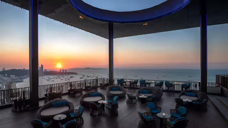 Rooftop lounge with modern seating overlooking the ocean at sunset, with a clear sky and distant city skyline, creating a peaceful, scenic atmosphere.