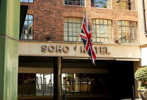 The entrance of the Soho Hotel, with a large Union Jack flag displayed above the doorway, set against a brick building with large windows.
