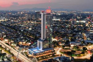 A tall, illuminated hotel building with a red RIU sign stands prominently in a cityscape at dusk, surrounded by roads, lights, and low-rise buildings under a colorful evening sky.
