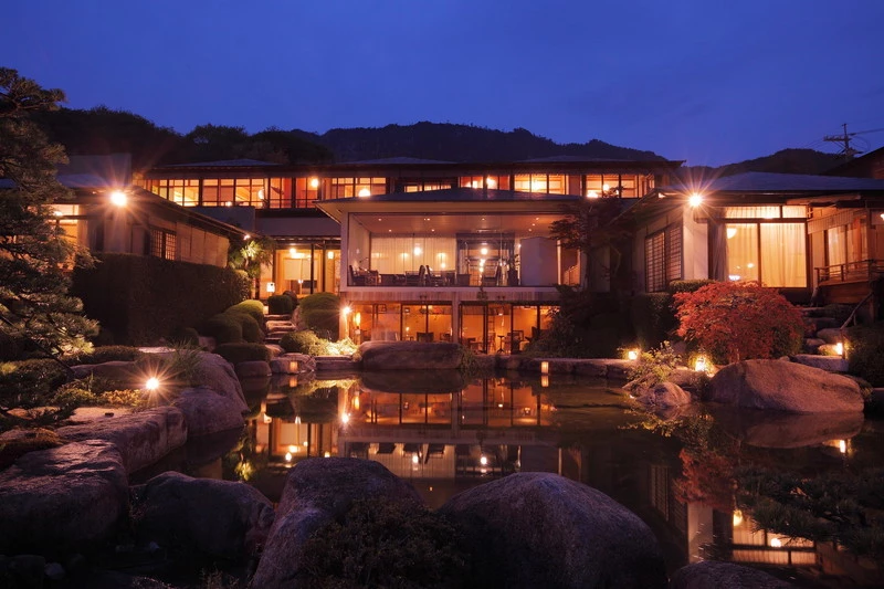 A traditional Japanese inn is warmly lit at night, with glowing windows reflecting in a tranquil garden pond surrounded by rocks and manicured greenery.