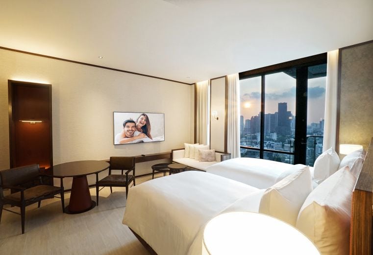 Modern hotel room with two large beds, a round table with chairs, a wall-mounted TV displaying a smiling couple, a sofa by the window, and a city skyline visible at sunset through floor-to-ceiling windows.