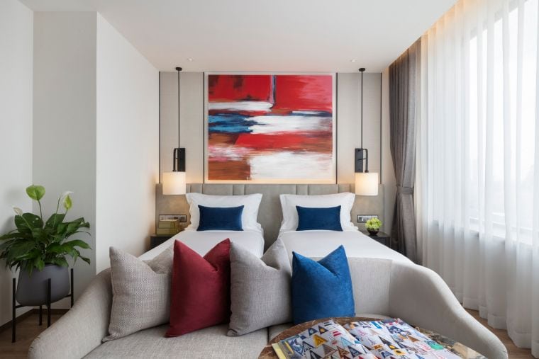 Modern hotel room with two single beds, blue and white pillows, a beige sofa with throw pillows, large abstract red and blue artwork above the beds, floor-to-ceiling window with sheer curtains, and a green potted plant.