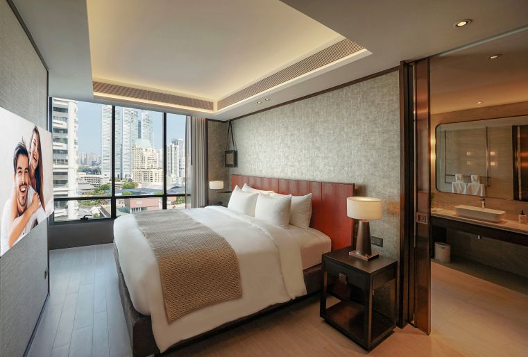 Modern hotel room with a large bed, bedside tables, a large window with a city view, a bathroom visible through a glass wall, and a TV screen showing a smiling couple on the left wall.