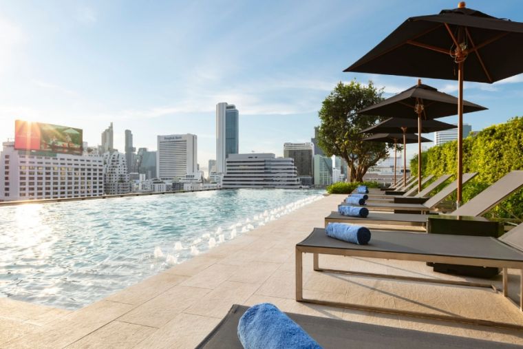 Rooftop infinity pool with lounge chairs and blue towels, shaded by umbrellas, overlooking a city skyline with tall buildings at sunset.