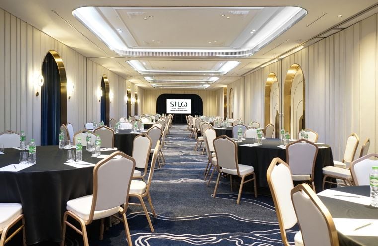 A modern conference room with round tables and white chairs arranged in rows. Water bottles and glasses are set on the tables. At the front, a screen displays SILQ. The room has elegant lighting and carpeted floors.
