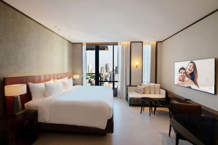 A modern hotel room with a large bed, bedside lamps, a seating area, a desk, and a wall-mounted TV displaying a smiling couple. Floor-to-ceiling windows offer a view of a city skyline.