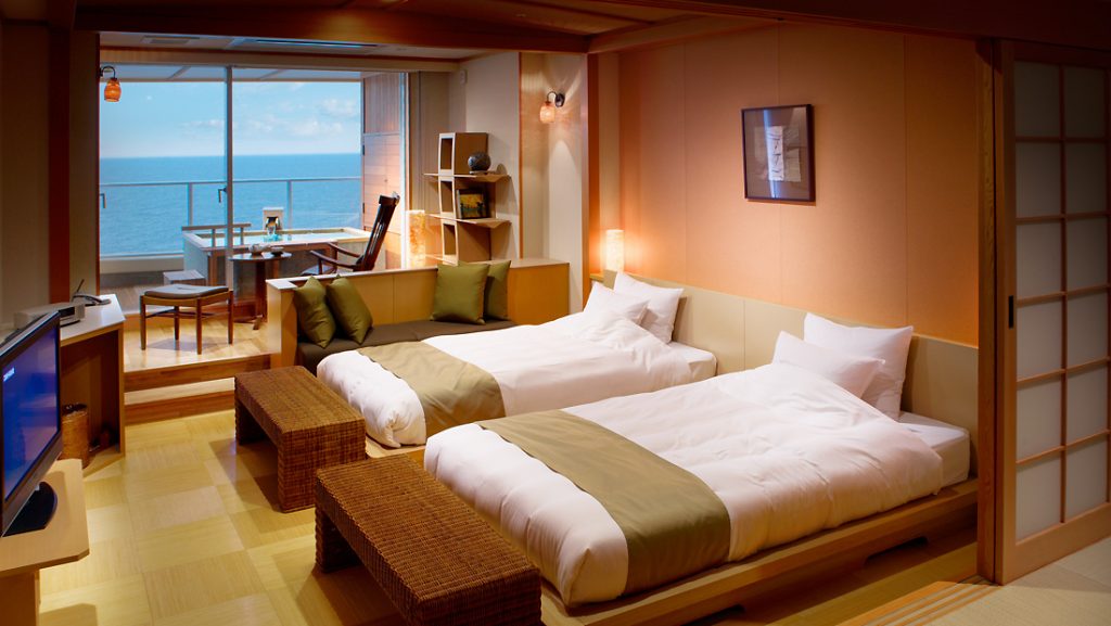 A modern Japanese-style hotel room with two twin beds, a small dining area by large windows overlooking the ocean, tatami flooring, and warm ambient lighting.