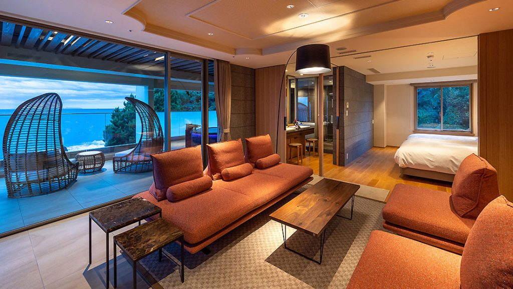 Modern hotel suite with orange couches, a wooden coffee table, and large windows showing an ocean view. The suite has a balcony with outdoor seating and a bedroom area visible through glass doors. Warm, ambient lighting throughout.