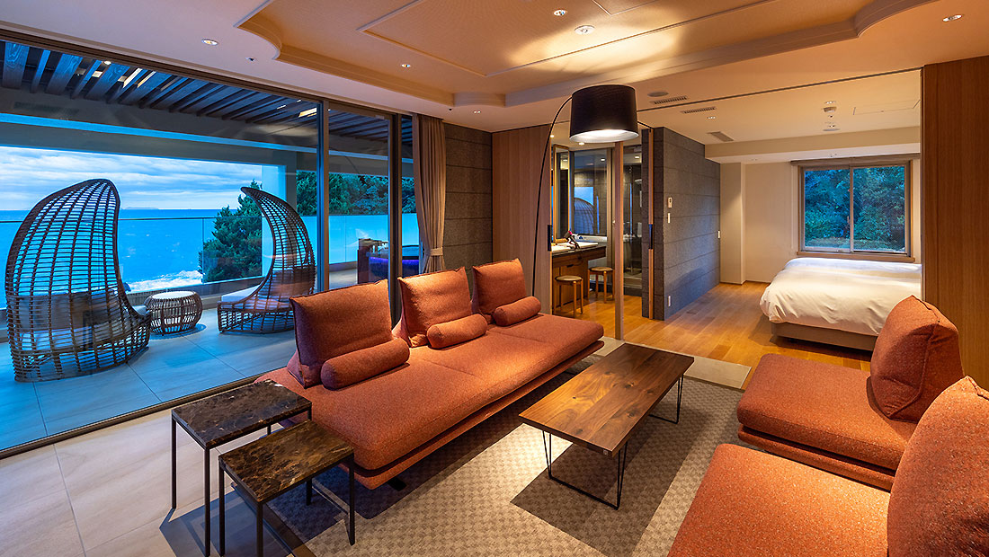 Modern hotel suite with orange couches, a wooden coffee table, and large windows showing an ocean view. The suite has a balcony with outdoor seating and a bedroom area visible through glass doors. Warm, ambient lighting throughout.