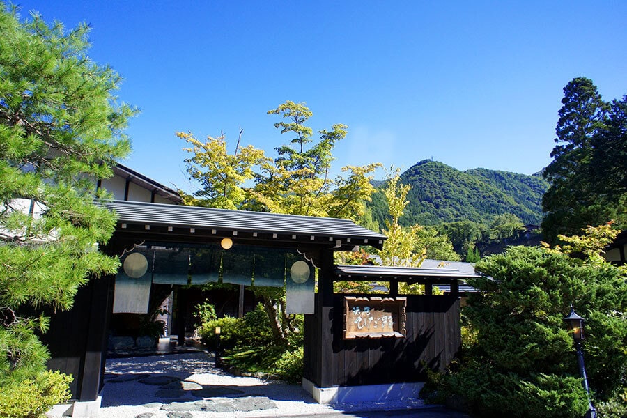 A traditional Japanese inn with a dark wooden entrance surrounded by lush greenery, set against a backdrop of forested mountains and a bright blue sky.