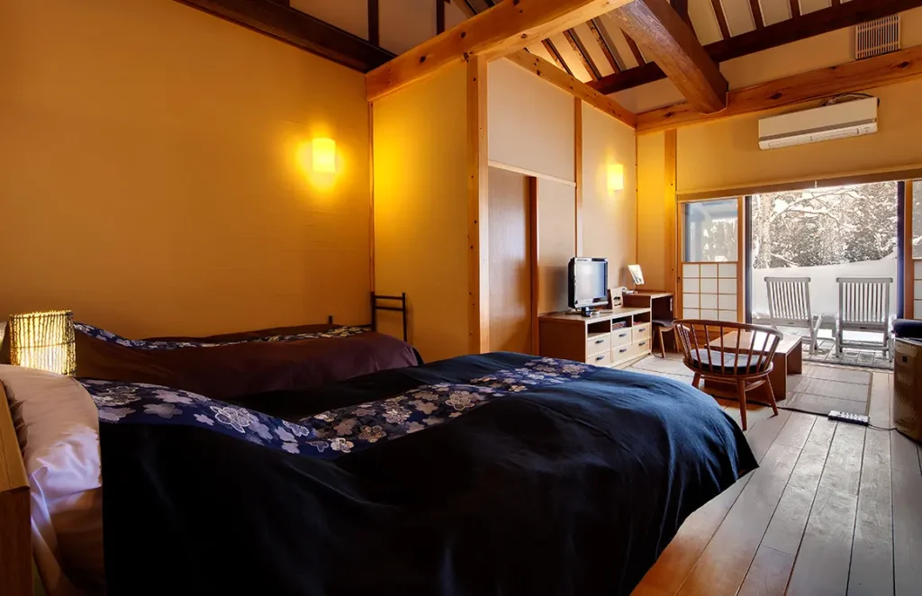 A cozy Japanese-style room with two beds, floral bed runners, wooden beams, warm lighting, a TV, a low table with chairs, and large windows opening to a balcony with outdoor seating and a scenic view.