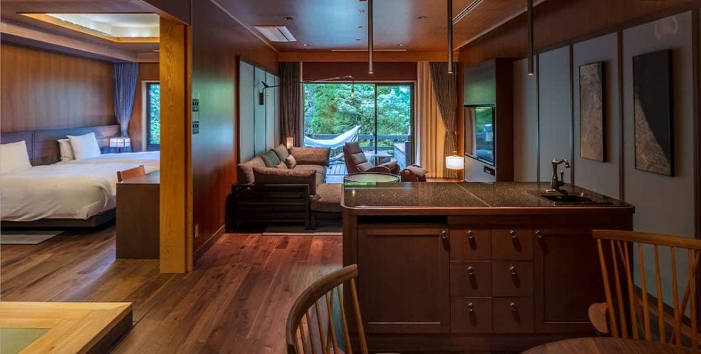 A spacious, modern hotel suite with wooden floors, a large bed, a living area with a sofa, kitchen island, and sliding glass doors leading to a balcony with a hammock and views of green trees outside.