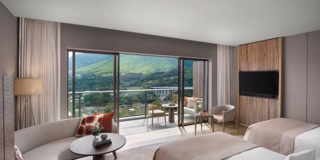 Modern hotel room with two beds, a sofa, a small round table, and a balcony overlooking lush green mountains and valley. Large sliding glass doors let in natural light, creating a bright and inviting space.