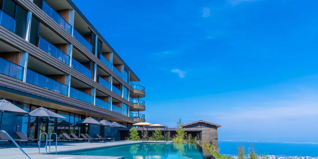 Modern hotel with balconies overlooks an infinity pool and lounge chairs with umbrellas, set against a bright blue sky and distant view of the sea.