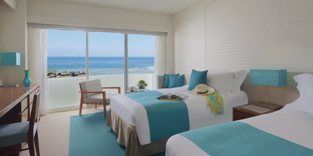 A bright hotel room with two beds, turquoise accents, a chair by the window, a desk, and a large sliding glass door offering an ocean view and letting in natural light.