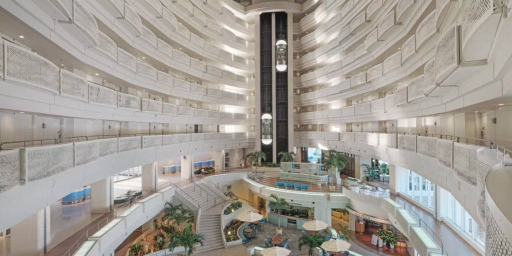 Spacious hotel atrium with multiple white balconies, tall central elevator, large windows, indoor plants, and seating areas with tables and umbrellas on the ground floor. Bright and modern architectural design.