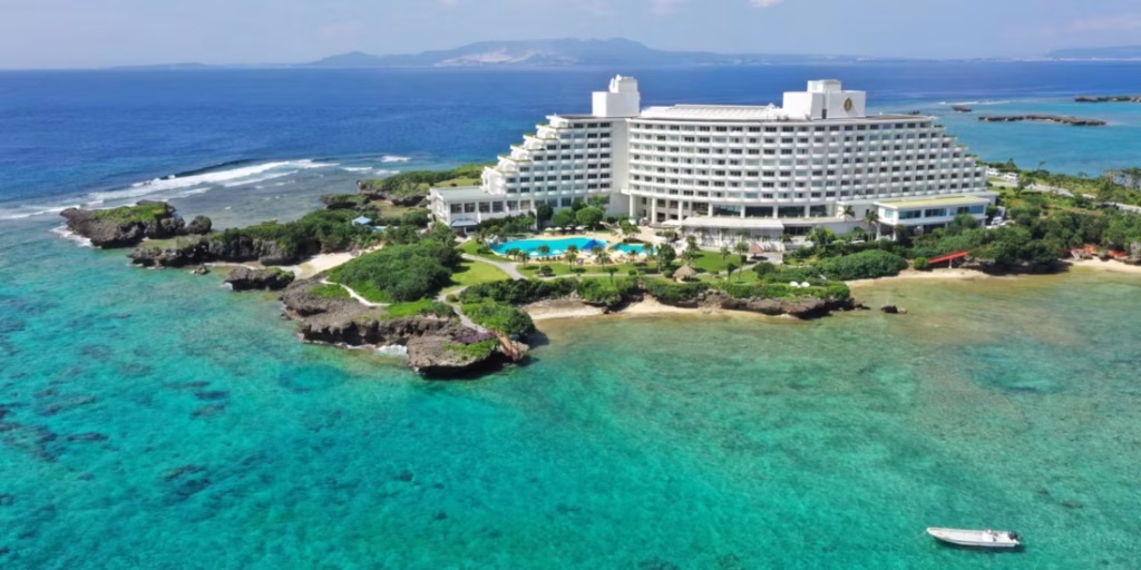 A large white hotel sits on a lush, rocky peninsula surrounded by clear turquoise ocean water, with a pool and gardens in front and a small boat in the foreground. An island is visible in the distance across the sea.