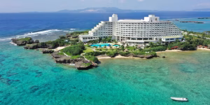 A large white hotel sits on a lush, rocky peninsula surrounded by clear turquoise ocean water, with a pool and gardens in front and a small boat in the foreground. An island is visible in the distance across the sea.